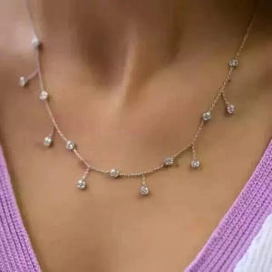 May include: A delicate gold necklace featuring multiple round, clear gemstones. The necklace has a fine chain with gemstones evenly spaced along the chain and dangling from it. The necklace is worn against a skin-toned background.