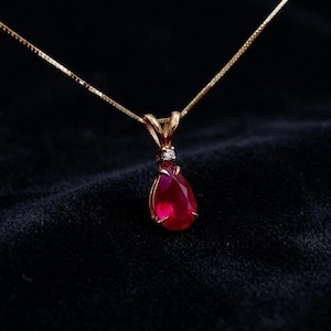 May include: A gold necklace featuring a pear-shaped red gemstone pendant suspended from a delicate chain. The pendant is accented with a single small clear stone set at the top, where the bail connects to the gold chain, creating a refined look.