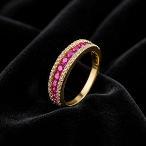 May include: A gold band ring featuring a central row of round, vibrant pink stones flanked by two thinner rows of small, clear white stones. The metal has a polished yellow finish, and the design showcases a channel setting for the pink stones.
