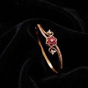 Puede incluir: Un brazalete dorado con un motivo central en forma de flor engastado con pequeñas piedras rojas y piedras blancas transparentes. La banda está acentuada con una hilera de diminutas piedras blancas transparentes a lo largo de la parte superior.