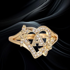 Peut inclure: Une bague dorée avec un motif en forme de cœur et la lettre "A" au centre. Le cœur est orné de petits diamants transparents. Des motifs floraux sont de chaque côté du cœur.