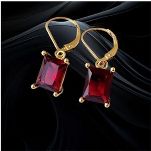 May include: A pair of gold-toned leverback earrings, each featuring a single rectangular-cut deep red gemstone held by four small prongs. The earrings have a polished metallic finish and a classic drop design, showcasing a vibrant crimson hue.