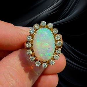 Könnte beinhalten: Ein goldener Ring mit einem ovalen Opal-Edelstein, umgeben von kleinen Diamanten. Der Opal zeigt ein Farbspiel mit blauen und grünen Farbtönen. Der Ring ist vor einem dunklen Hintergrund platziert.