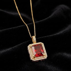 May include: A gold necklace featuring a rectangular pendant with a large, deep red gemstone at the center. The pendant is framed by two rows of small, clear white stones, and the bail is also encrusted with tiny clear stones on a gold chain.
