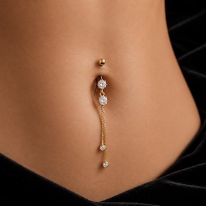 May include: A gold-toned belly button ring featuring a top ball, a larger round clear stone, and a second round clear stone connected to two delicate gold chains. Each chain terminates in a small, round clear stone, creating a dangling effect.