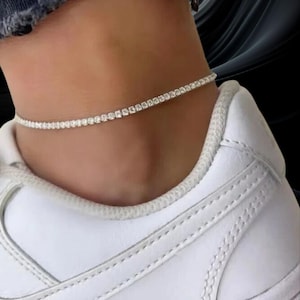 May include: A delicate silver anklet with small, sparkling stones encircles an ankle. The anklet is worn above a white sneaker, creating a contrast between casual and elegant styles. The stones appear to be evenly spaced and catch the light.