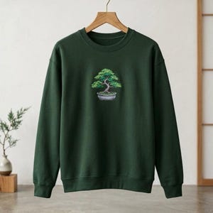 May include: Dark green sweatshirt featuring an embroidered bonsai tree design. The bonsai tree has green leaves and a brown trunk, planted in a gray pot. The sweatshirt is displayed on a wooden hanger.