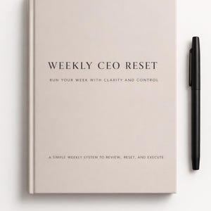 May include: A light beige hardcover notebook with the text "WEEKLY CEO RESET" and the phrases "RUN YOUR WEEK WITH CLARITY AND CONTROL" and "A SIMPLE WEEKLY SYSTEM TO REVIEW, RESET, AND EXECUTE". A black pen rests beside the notebook.