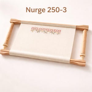 May include: A wooden embroidery hoop with a cream-colored fabric stretched across it. The fabric features a floral cross-stitch design in shades of pink, yellow, and green. The text "Nurge 250-3" is printed at the top.