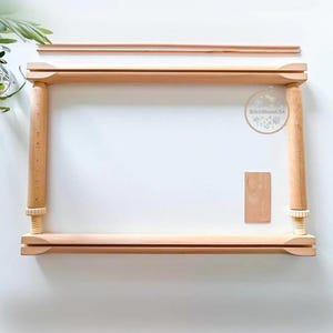 May include: A wooden embroidery hoop frame, featuring a rectangular design with rounded corner posts and adjustable screw knobs. The frame is made of light-colored wood and includes a small wooden piece. The frame is designed for needlework and embroidery projects.