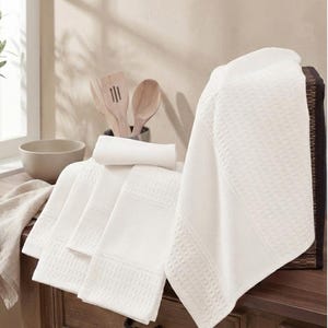 May include: A collection of white waffle-weave kitchen towels, displayed on a wooden surface. The towels feature a textured design and are folded, with one towel draped over the edge. Wooden kitchen utensils and a bowl are in the background.