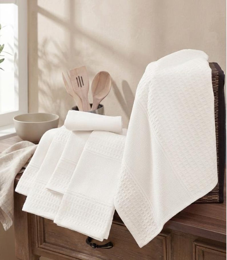May include: A collection of white waffle-weave kitchen towels, displayed on a wooden surface. The towels feature a textured design and are folded, with one towel draped over the edge. Wooden kitchen utensils and a bowl are in the background.