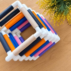 May include: A stack of colorful embroidery hoops with white plastic frames and cushioned grips in various colors, including navy, orange, pink, and beige. The brand name "Nurge" is printed on each grip. A sprig of yellow flowers is in the background.