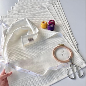 May include: Cream waffle-weave towels with a decorative ribbon, embroidery floss, embroidery hoop, and scissors. The towels have a textured pattern and a label that reads "SARRA HOME". The embroidery floss is yellow, purple, and magenta.