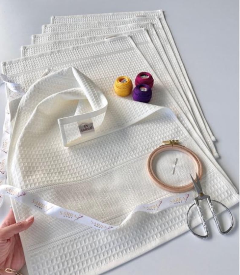 May include: Cream waffle-weave towels with a decorative ribbon, embroidery floss, embroidery hoop, and scissors. The towels have a textured pattern and a label that reads "SARRA HOME". The embroidery floss is yellow, purple, and magenta.