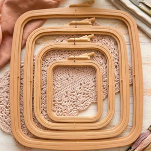 May include: Four square-shaped embroidery hoops in varying sizes, stacked on top of each other. The hoops are a light brown color and have a tightening mechanism at the top. The sizes are labeled with measurements in millimeters.