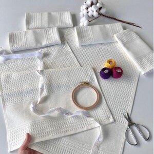 May include: A collection of white waffle-weave towels and flannels, some folded, with patterned edges. There are also yellow, purple, and pink embroidery threads, a wooden embroidery hoop, and scissors. A sprig of cotton adds a natural element to the scene.