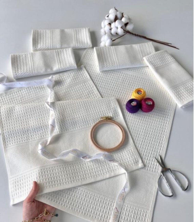 May include: A collection of white waffle-weave towels and flannels, some folded, with patterned edges. There are also yellow, purple, and pink embroidery threads, a wooden embroidery hoop, and scissors. A sprig of cotton adds a natural element to the scene.