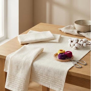 May include: A collection of cream-coloured waffle-weave kitchen towels and a table runner are displayed on a wooden table. A small wooden tray holds colourful spools of thread, and scissors are placed nearby. A sprig of cotton and a ceramic bowl are also present.