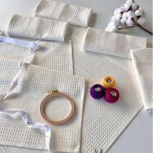 May include: A collection of white waffle-weave towels, an embroidery hoop, and colourful thread spools. A sprig of cotton and scissors are also visible. The items are arranged on a white surface, suggesting a craft or home decor theme.
