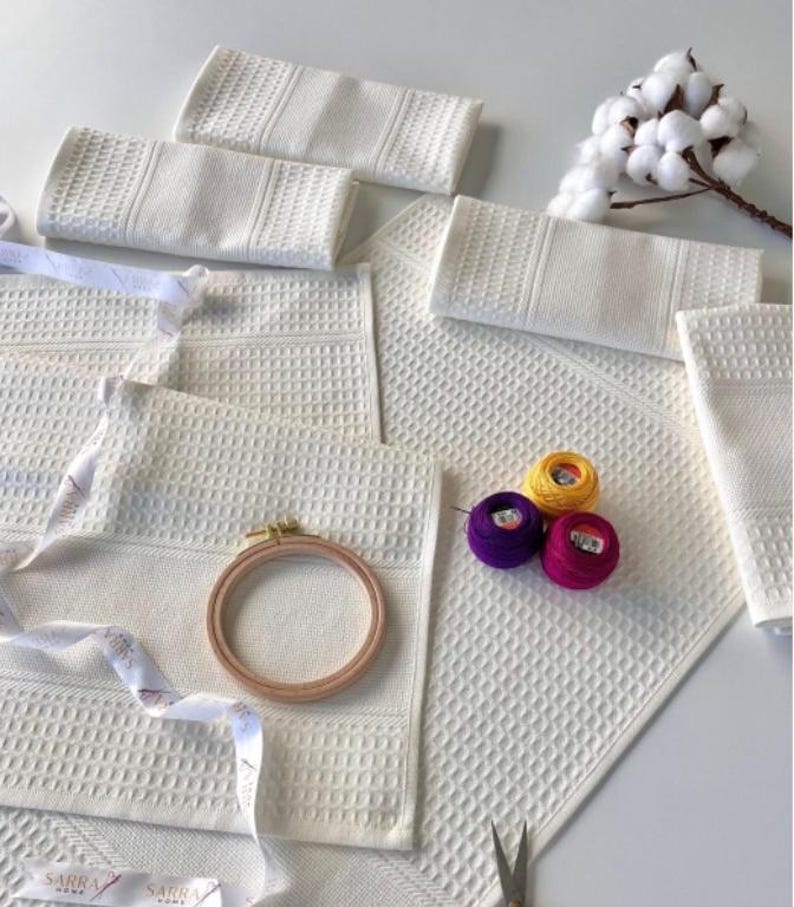 May include: A collection of white waffle-weave towels, an embroidery hoop, and colourful thread spools. A sprig of cotton and scissors are also visible. The items are arranged on a white surface, suggesting a craft or home decor theme.