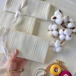 May include: Off-white waffle-weave towels with a textured pattern. A white ribbon with text is draped across the towels. Cotton sprigs and embroidery supplies are also present, suggesting a home decor or textile theme.