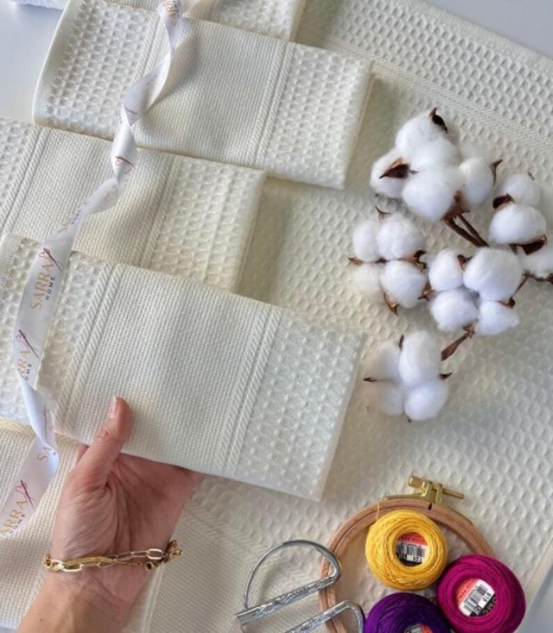 May include: Off-white waffle-weave towels with a textured pattern. A white ribbon with text is draped across the towels. Cotton sprigs and embroidery supplies are also present, suggesting a home decor or textile theme.