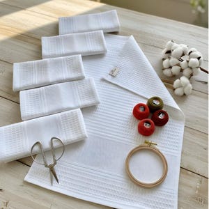 May include: A collection of white waffle-weave kitchen towels, a larger matching towel, and several small spools of thread in red and green. A wooden embroidery hoop and vintage scissors are also present, suggesting a crafting theme.