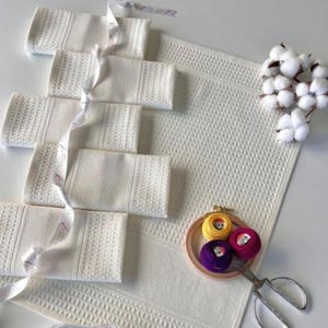 May include: White waffle-weave towels, folded and tied with ribbon, and a matching bath mat. Embroidery threads in yellow, pink, and purple are in a wooden hoop with scissors. Cotton sprigs provide a natural accent.