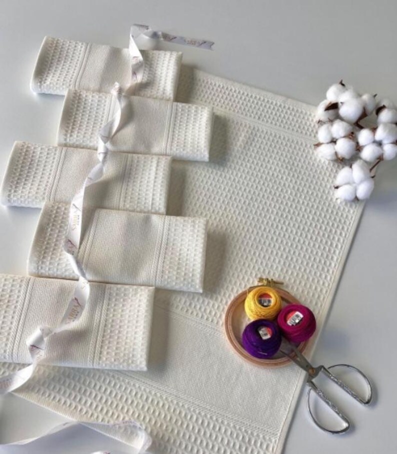 May include: White waffle-weave towels, folded and tied with ribbon, and a matching bath mat. Embroidery threads in yellow, pink, and purple are in a wooden hoop with scissors. Cotton sprigs provide a natural accent.