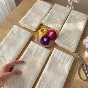 May include: Cream-coloured waffle-weave towels arranged on a wooden surface. A small wooden embroidery hoop holds yellow, pink, and purple thread spools. Scissors and cotton bolls are also visible.