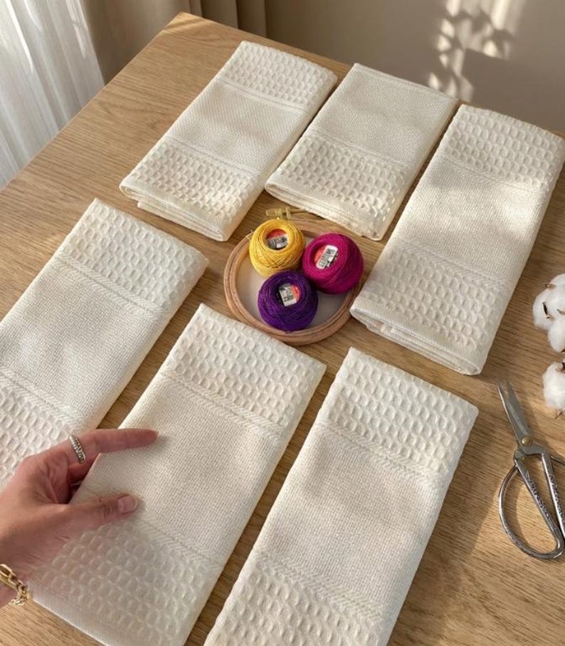 May include: Cream-coloured waffle-weave towels arranged on a wooden surface. A small wooden embroidery hoop holds yellow, pink, and purple thread spools. Scissors and cotton bolls are also visible.