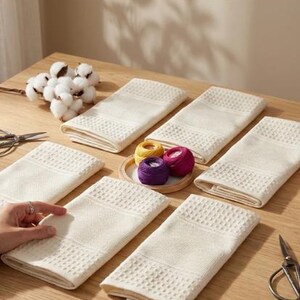 May include: Several cream-coloured, folded rectangular towels with a textured pattern are arranged on a wooden table. A small wooden tray holds spools of yellow, purple, and pink thread. Cotton bolls and scissors are also visible.