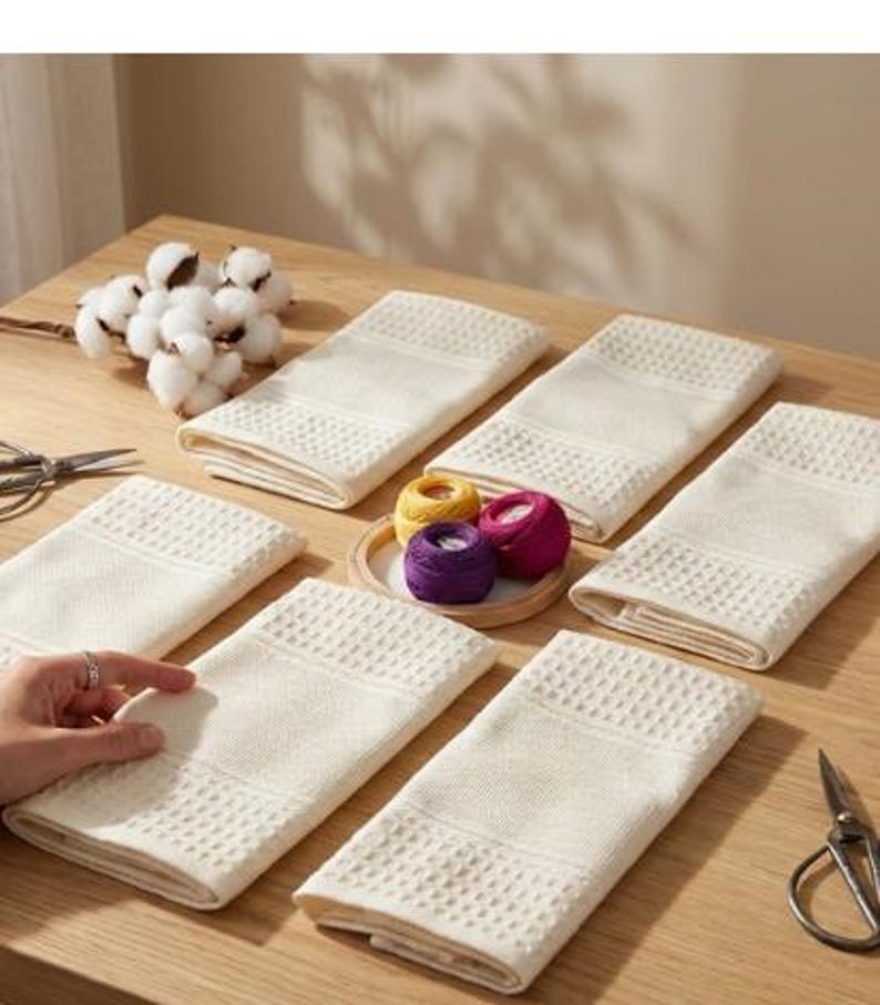 May include: Several cream-coloured, folded rectangular towels with a textured pattern are arranged on a wooden table. A small wooden tray holds spools of yellow, purple, and pink thread. Cotton bolls and scissors are also visible.