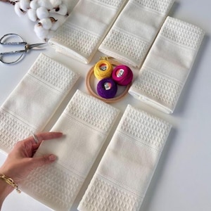 May include: Six white waffle-weave towels with textured borders are displayed. The towels are arranged on a white surface, with an embroidery hoop holding colourful thread spools, cotton sprigs, and a pair of scissors.