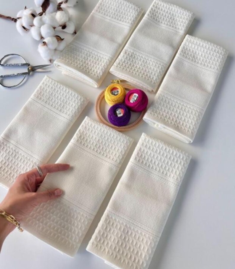 May include: Six white waffle-weave towels with textured borders are displayed. The towels are arranged on a white surface, with an embroidery hoop holding colourful thread spools, cotton sprigs, and a pair of scissors.