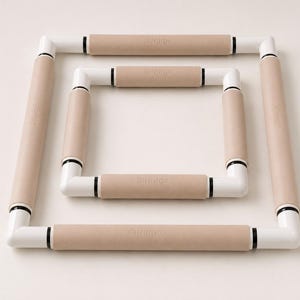 May include: Two square embroidery frames nested inside each other. Each frame consists of white plastic corner joints connected by light tan cylindrical bars. The word Nurge is embossed on each of the eight tan bars of the two frames.