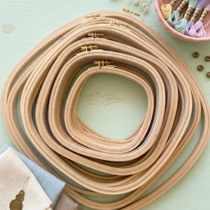 May include: A collection of square wooden embroidery hoops in various sizes, stacked together. The hoops are light brown with brass hardware. Embroidery floss in pastel colors and fabric samples are also visible, suggesting a crafting theme.