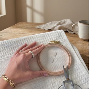 May include: A wooden embroidery hoop with white fabric stretched inside, held over a white waffle-weave towel. Small scissors and a gold bracelet are also visible. The scene is set on a wooden table with a neutral background.
