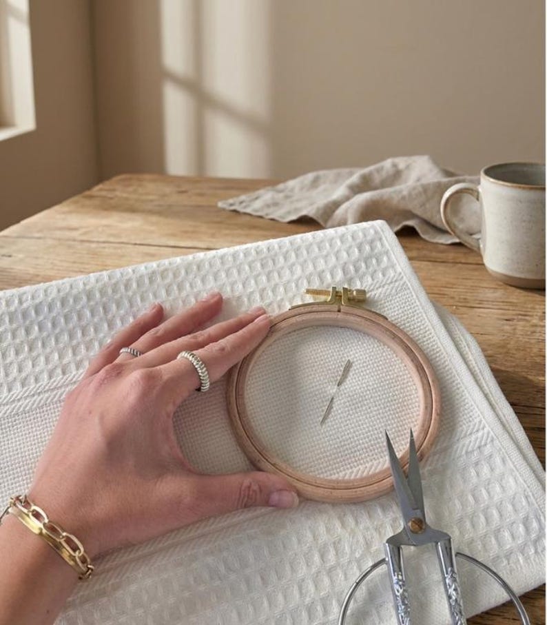 May include: A wooden embroidery hoop with white fabric stretched inside, held over a white waffle-weave towel. Small scissors and a gold bracelet are also visible. The scene is set on a wooden table with a neutral background.