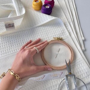 May include: White waffle-weave towels with a SARRA label. An embroidery hoop, scissors, and spools of yellow, purple, and pink thread are visible. A hand wearing rings and a gold bracelet holds the embroidery hoop.