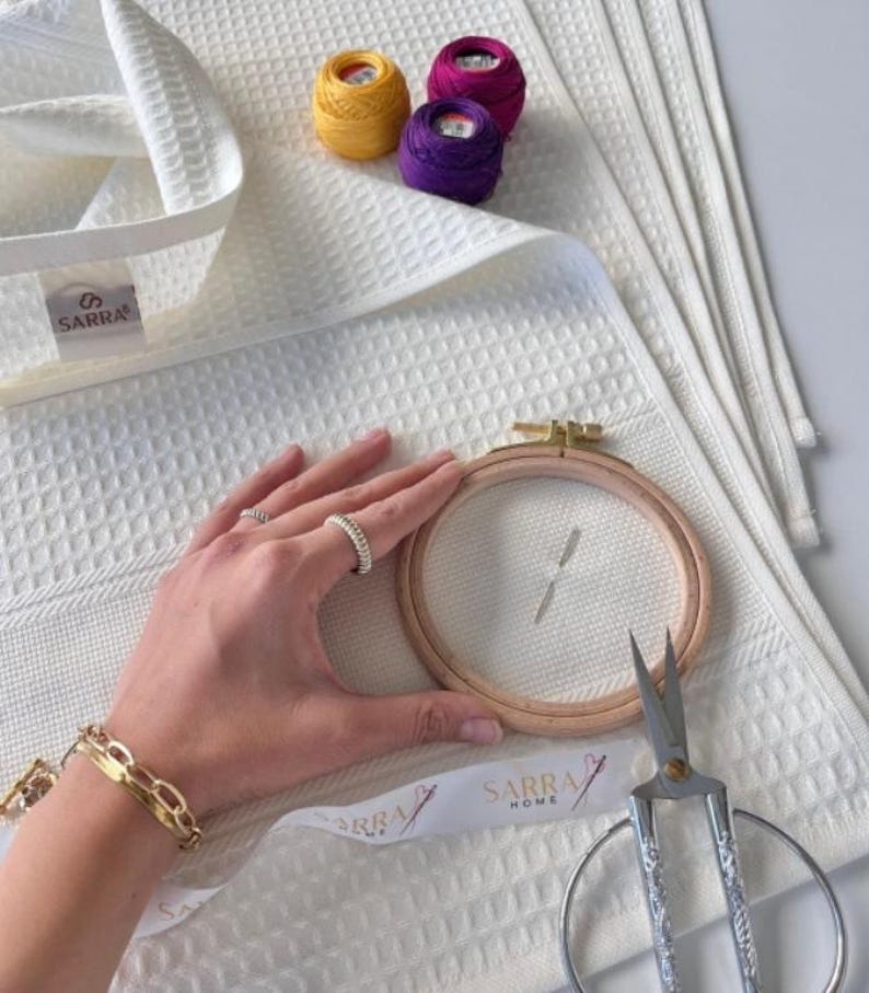 May include: White waffle-weave towels with a SARRA label. An embroidery hoop, scissors, and spools of yellow, purple, and pink thread are visible. A hand wearing rings and a gold bracelet holds the embroidery hoop.