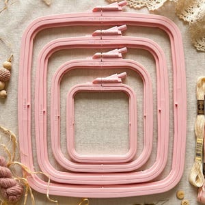 May include: A set of four pink square embroidery hoops of varying sizes, with measurements in millimeters. The largest hoop is labeled "No. 4 - 280 mm". The hoops are arranged in a nested fashion, with a screw mechanism at the top. The image includes embroidery floss and other craft supplies.