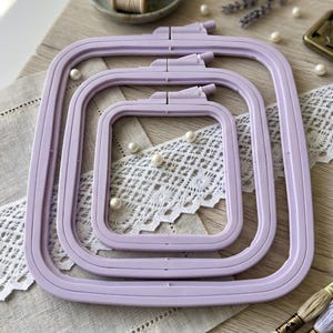 May include: Three square, lavender-colored embroidery hoops of varying sizes are nested together. The hoops are made of plastic and have a tightening mechanism at the top. The background includes lace, pearls, and spools of thread, suggesting a crafting theme.