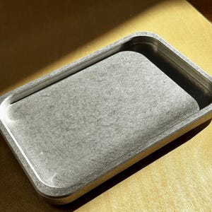 May include: A rectangular metal tray with a brushed silver finish and rounded corners. The tray features a flat base with slightly raised, smooth edges, showcasing a metallic texture and a consistent industrial appearance throughout the surface.