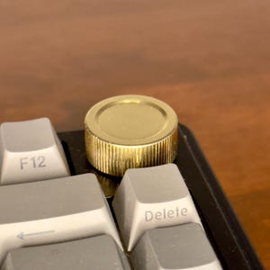 May include: A circular gold-colored metal knob with a textured, ridged side edge, positioned on a black computer keyboard base next to light gray keycaps labeled F12, Delete, and PgUp. The knob features a smooth, flat top surface.