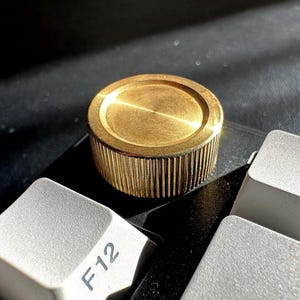 May include: A gold-colored, cylindrical object with a ridged side and a flat top, resting on a keyboard. The keycap of the F12 key is visible. The object's design suggests a mechanical component or decorative element.
