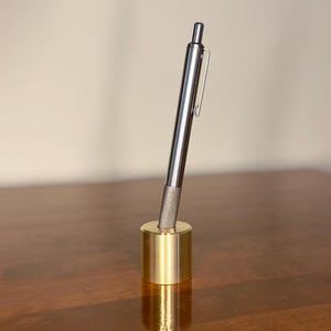 May include: A metallic silver pen with a textured grip and a pocket clip, standing upright in a cylindrical, polished gold-toned brass pen holder. The pen is positioned at a slight angle within the circular opening of the heavy metal base.