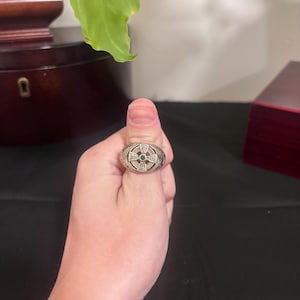 Puede incluir: Un anillo de tono plateado con una cara circular que presenta un diseño de cruz celta en relieve. El centro de la cruz está acentuado con una pequeña piedra redonda de color verde. La banda y el área alrededor de la cruz muestran metal.