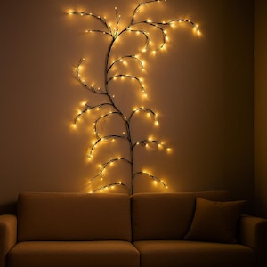 Puede incluir: Una guirnalda de luces decorativa montada en la pared con forma de rama de árbol, con luces amarillas cálidas. La lámpara está montada en una pared de color neutro sobre un sofá beige con un cojín a juego. Las luces crean un ambiente acogedor.